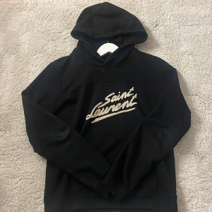 Svart hoodie från Saint Laurent - Säljer en stilren svart hoodie från Saint Laurent med deras ikoniska logga i vitt på bröstet. Perfekt för en casual look med en touch av lyx. Hoodien har en klassisk passform och är tillverkad i mjukt material för extra komfort.  Jag har inget bevis på att tröjan är äkta därav priset.