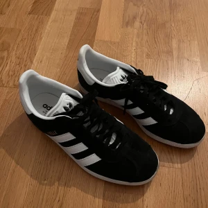 Svarta Adidas Gazelle sneakers - Snygga svarta Adidas Gazelle sneakers med vita detaljer och klassiska tre ränder på sidan. Skorna har snörning och en vit sula som ger en stilren look. Perfekta för en casual stil. använda 2 gånger 