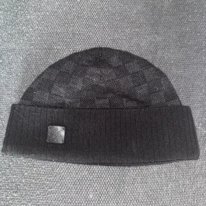 Svart mössa från Louis Vuitton - Elegant svart mössa från Louis Vuitton med diskret rutmönster och ribbad kant. Tillverkad i Italien, perfekt för en stilren look. Märkesdetalj framtill ger en exklusiv touch.