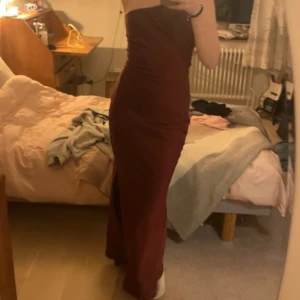 Vinröd långklänning - Säljer en elegant vinröd långklänning med en stilren assymetrisk oneshoulder-design och smala axelband. Klänningen har en slits som ger en extra touch av stil😍. Perfekt för speciella tillfällen som t.ex bal eller liknande💕Klänningen är i satin material
