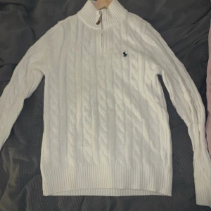 Vit stickad tröja från Ralph Lauren - Säljer en stilren vit stickad tröja från Ralph Lauren med dragkedja och klassiskt kabelstickat mönster. Tröjan har en broderad logga på bröstet och ribbade muddar. Perfekt för kyliga dagar!