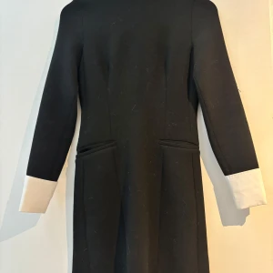 Svart klänning Zara - Elegant svart klänning med vita ärmslut. Den har en stilren design med långa ärmar och en klassisk passform. Perfekt för en chic look.