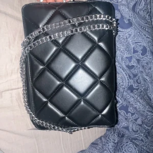 Svart quiltad axelväska med kedja - Snygg svart axelväska i quiltat mönster med silverfärgad kedja som axelrem aldrig använd. Väskan har en dragkedja för säker förvaring och en stilren design som passar perfekt för en chic look.