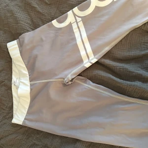 Grå leggings med vit text - Snygga grå leggings med vit text längs benet. De har en elastisk midja och är tillverkade i ett stretchigt material som ger en tight passform. Perfekta för en sportig look.