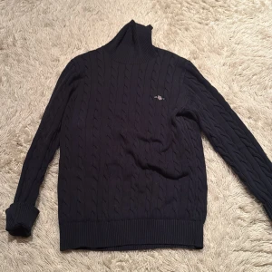 Mörkblå kabelstickad polotröja från Gant - Snygg mörkblå kabelstickad polotröja från Gant med ribbad krage och muddar. Perfekt för kyliga dagar med sin stilrena design och varma material. Helt ny