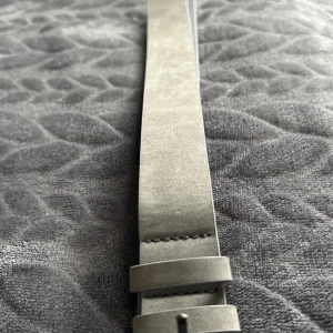 Grått bälte i mocka - Snyggt grått bälte i mocka med en stilren design. Perfekt för att ge en elegant touch till din outfit. Bältet har en enkel spänne och passar bra till både jeans och kostymbyxor.