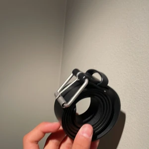 Svart skärp med silverfärgat spänne - Snyggt svart skärp i skinn med ett stilrent silverfärgat spänne. Perfekt för att ge en klassisk touch till din outfit. Skärpet har flera hål för justerbar passform.