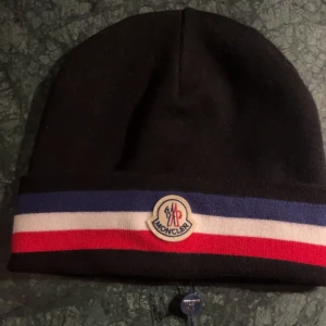 Svart mössa från Moncler - Snygg svart mössa från Moncler med röd, vit och blå randdetalj. Perfekt för att hålla värmen med stil. Märkeslogga framtill ger en exklusiv touch.