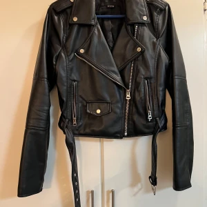 Svart skinnjacka från Pull&Bear - Snygg svart skinnjacka från Pull&Bear med dragkedjor och bälte. Jackan har en klassisk bikerstil med asymmetrisk dragkedja och knappar på kragen. Perfekt för en cool look!