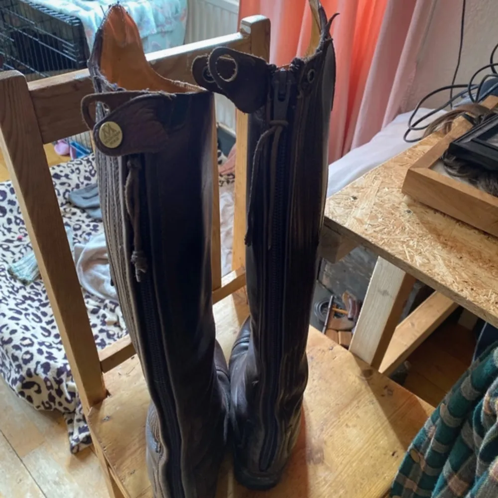 Säljer ett par bruna ridstövlar i skinn med dragkedja baktill. Stövlarna har en klassisk design med rundad tå och dekorativa knappar upptill. Perfekta för ridning eller en stilren look. Aurora . Kengät.