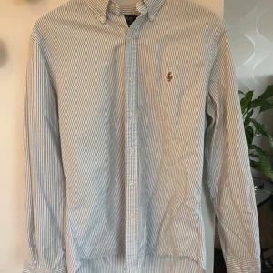 Randig skjorta från Ralph Lauren - Snygg och klassisk långärmad skjorta från Ralph Lauren i blått och vitt randigt mönster. Skjortan har knappar framtill och en broderad logga på bröstet. Perfekt för en stilren look!