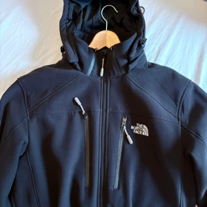 Svart jacka från The North Face - Säljer en svart jacka från The North Face med dragkedja och flera praktiska fickor. Jackan har en huva och ett märke på ärmen. Perfekt för kyligare dagar och outdoor-äventyr.