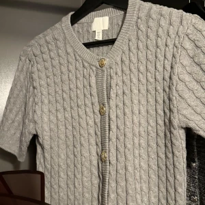 Grå stickad kofta från H&M - Säljer en stilren grå stickad kofta från H&M med korta ärmar och snygga guldknappar framtill. Perfekt för lager-på-lager och att hålla sig varm under kyligare dagar. Passar både till jeans och kjol. 🧥✨