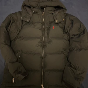 Svart dunjacka från Ralph Lauren - Säljer en snygg svart dunjacka från Ralph Lauren med en klassisk design. Jackan har en dragkedja framtill och en liten röd logotyp på bröstet. Perfekt för kyliga dagar med sin varma och bekväma passform.