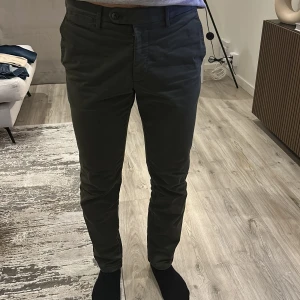 Mörkgrå chinos från Oscar Jacobsson - Snygga mörkgrå chinos med normal passform. Byxorna har en klassisk design med knappar och dragkedja framtill. Perfekta för en stilren look. Storlek 46