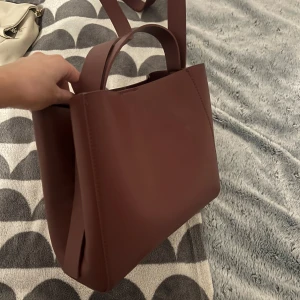 Vinröd väska gina tricot - Snygg vindöd axelväska i syntetmaterial med stilren design. Väskan har en praktisk dragkedja och två handtag för enkel bärning. Perfekt för att bära med sig det nödvändigaste med stil. Från gina helt oanvänd🩷