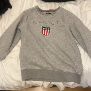 Grå sweatshirt från GANT - Säljer en stilren grå sweatshirt från GANT med broderad logga och ett emblem på bröstet. Tröjan har långa ärmar och en rund halsringning med ribbade kanter. Perfekt för en avslappnad stil.