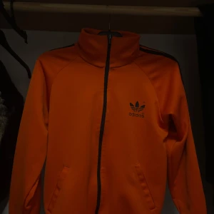 Tröja från adidas - Säljer en cool orange tröja från Adidas med svart logga och dragkedja. Tröjan har långa ärmar och svarta ränder längs ärmarna. Perfekt för en sportig look!