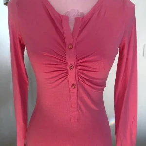 Rosa långärmad topp från Ilse Jacobsen - Säljer en snygg rosa långärmad topp från Ilse Jacobsen. Toppen har en v-ringad design med knappar framtill och en smickrande rynkdetalj vid bysten. Perfekt för en stilren look!