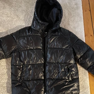 Svart pufferjacka från lager 157 - Säljer en svart pufferjacka från lager 157. Jackan har en glansig yta och är utrustad med en dragkedja framtill. Perfekt för kyliga dagar med sin vadderade design och huva.