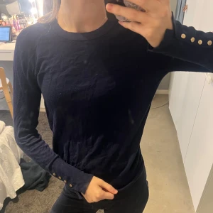 Mörkblå tröja från Zara - Säljer en stilren mörkblå tröja från Zara med långa ärmar och dekorativa knappar vid ärmsluten. Perfekt för en elegant och avslappnad look. Storleken är M men på mig som brukar ha xs/s passar den väldigt bra.