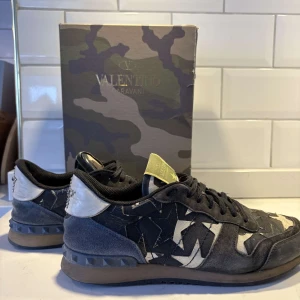 Valentino Rockrunner sneakers i kamouflage - Snygga Valentino Rockrunner sneakers med ett coolt kamouflagemönster i svart, grått och vitt. Skorna har snörning och en unik design med stjärndetaljer. Perfekta för att ge din outfit en edgy touch.