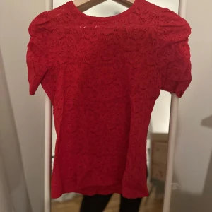 Röd spetsblus från Kapphal - Säljer en röd spetsblus med korta ärmar och ett elegant mönster. Perfekt för att lägga till en färgklick i garderoben. Blusen har en rund halsringning och en feminin touch med sina puffiga ärmar.