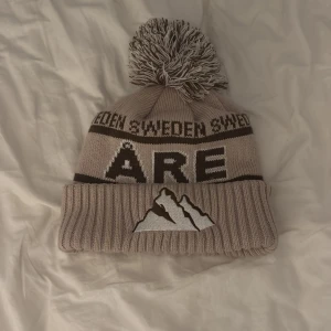 Beige mössa med tofs från Åre  - Snygg beige mössa med stor tofs och texten 'Åre' samt bergsmotiv. Mössan har en ribbad kant och texten 'Sweden' runt om. Perfekt för kalla dagar. Pirs går att diskuteras 
