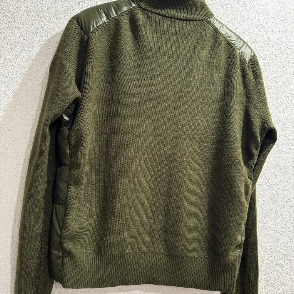 Snygg olivgrön dunjacka från Moncler med stickade ärmar och quiltad front. Jackan har en dragkedja med röd och vit kant, samt Moncler-logga på ärmen. Perfekt för kyliga dagar med stil.. Takit.