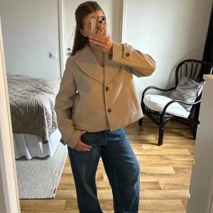 Beige kappa från Vila - Säljer en stilren beige kappa från Vila med stora slag och knappar framtill. Den är aldrig använd men har ingen lapp kvar. Storlek 38. Jackan har en klassisk design med långa ärmar och fickor på sidorna. 