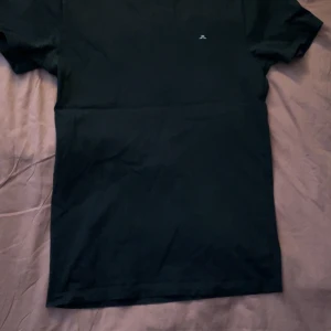 Svart t-shirt från J.Lindeberg - Säljer en stilren svart t-shirt från J.Lindeberg. Den har en liten logga på bröstet och är tillverkad i mjukt material för en bekväm passform. Perfekt för en enkel och snygg look. Skriv vid fundering✅