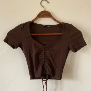 Brun ribbad topp från Zara - Säljer en snygg brun ribbad topp från Zara med korta ärmar och knytning framtill. Toppen har en croppad passform och en v-ringad halslinje. Perfekt för en trendig look!
