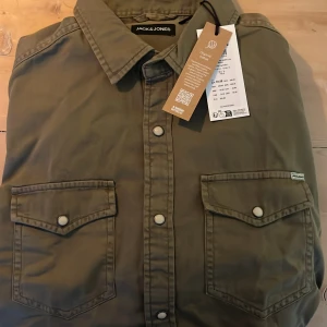 Olivgrön skjorta från Jack & Jones - Snygg olivgrön skjorta från Jack & Jones med två bröstfickor och knappar framtill. Perfekt för en avslappnad stil. Skjortan har långa ärmar och en klassisk krage.