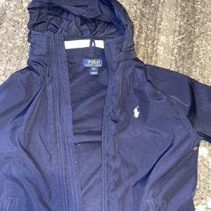 Mörkblå vindjacka från Polo Ralph Lauren - Snygg mörkblå vindjacka från Polo Ralph Lauren med dragkedja och huva. Jackan har en klassisk design med det ikoniska logotypen på bröstet. Perfekt för blåsiga dagar.