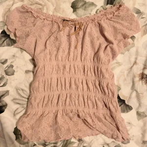 Rosa spetsblus från New Look - Söt rosa omlott spetsblus från New Look med korta off-shoulder ärmar och volangdetaljer, spets detaljer. Blusen har en liten knytning framtill och en elastisk smickrande passform. 