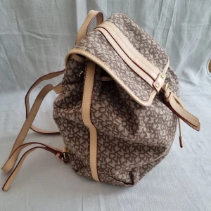 Brun mönstrad ryggsäck med läderdetaljer - DKNY  - Brun ryggsäck med DKNY monogrammönster och ljusa läderdetaljer. Väskan har justerbara axelremmar och en praktisk stängning med både spänne och snörning. Perfekt  storlek och 90 tals stil. Äkta. 