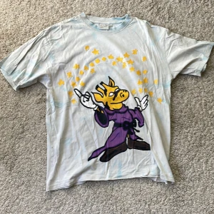 Vit t-shirt med tryck från Yoke - Säljer en vit t-shirt från Yoke med ett coolt tryck av en giraff liknande figur i lila kläder och gula stjärnor. T-shirten har korta ärmar och är i en normal passform. Perfekt för dig som gillar unika motiv! Den är i fint skick och säljer den för att jag inte använder den längre. Den har dock några enstaka hål i sig längst ner (se bilden ovan).