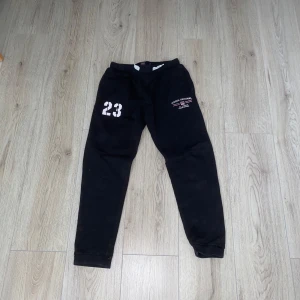 Svarta mjukisbyxor  - Säljer ett par svarta mjukisbyxor från Vinson Original med trycket '23' på ena benet och logotypen på det andra. Byxorna har en elastisk midja med snörning för justerbar passform. Perfekta för en avslappnad stil.