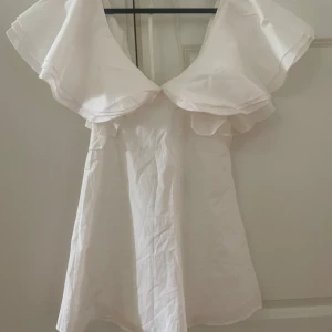 Vit klänning från Thanne - Säljer en söt vit klänning från Thanne med volangdetaljer och v-ringad design. Klänningen har en kort längd och är perfekt för sommarens varma dagar. Den är ärmlös och har en luftig passform som ger en romantisk känsla.