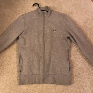 Grå tröja med dragkedja från Lacoste - Säljer en stilren grå tröja från Lacoste med dragkedja och det klassiska krokodilmärket på bröstet. Tröjan har långa ärmar och ribbade muddar vid ärmslut och nederkant. Perfekt för en avslappnad look.