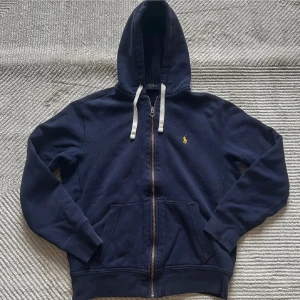 Mörkblå hoodie från Polo Ralph Lauren - Säljer en snygg mörkblå hoodie från Polo Ralph Lauren med dragkedja och huva. Den har vita snören och det klassiska gula logotypbroderiet på bröstet. Perfekt för en avslappnad stil.