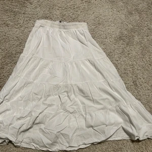 Vit långkjol från Fiore Fatto - Säljer en vit långkjol från Fiore Fatto med dragkedja som man kan ha fram eller bak. Kjolen har flera lager som ger en fin volangeffekt. Perfekt för en somrig look!