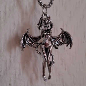 Silverfärgat halsband med demonhänge - Unikt silverfärgat halsband med ett detaljerat demonhänge. Hänget föreställer en demon med vingar och svans, vilket ger ett mystiskt och edgy intryck. Perfekt för dig som vill ha något annorlunda och iögonfallande.