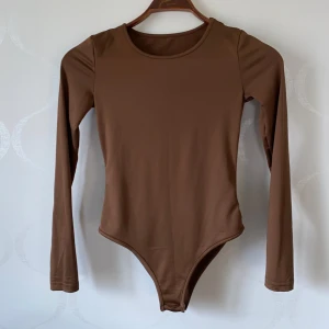 Brun långärmad bodysuit - Säljer en stilren brun långärmad bodysuit med rund halsringning. Perfekt för lager-på-lager eller att bära som den är. Den har praktiska knappar i grenen för enkel på- och avtagning.