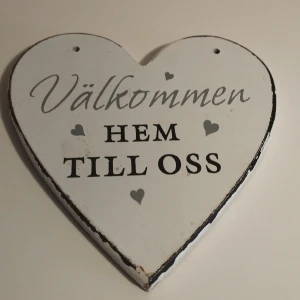 Hjärtaformad skylt med texten 'Välkommen hem till oss' - Vitt charmig hjärtaformad skylt i trä med texten 'Välkommen hem till oss' i svart och grått. Skylten har små hjärtan som dekoration och är perfekt för att ge en välkomnande känsla i hemmet. Den har en rustik look med slitna kanter. Storlek 12x12 cm.