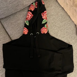 Croppad hoodie med broderade blomtryck - Säljer en svart hoodie från med färgglada blommor broderade på huvan och vida ärmar. Hoodien har en klassisk passform med dragsko i huvan.