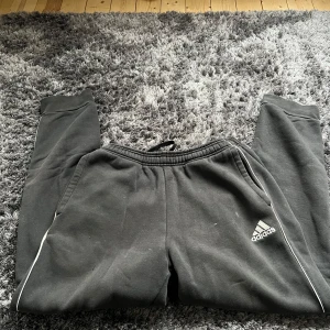 Svarta mjukisbyxor från Adidas - Säljer ett par svarta mjukisbyxor från Adidas med vita detaljer. Byxorna har en elastisk midja och är perfekta för en avslappnad stil. Adidas-loggan syns på ena benet.