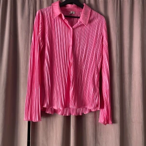 Rosa plisserad skjorta - Snygg rosa skjorta med plisserat mönster och knappar framtill. Den har en klassisk krage och långa ärmar, perfekt för en stilren look. Passar bra till både jeans och kjol för en chic outfit.
