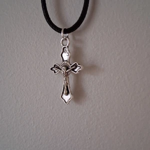 Silverfärgat korshalsband krucifix  - Snyggt silverfärgat korshalsband med detaljerad design. Hänget har en klassisk korsform med en figur i mitten och hänger på en svart snodd. Perfekt för att ge en stilren touch till din outfit.
