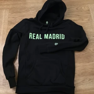 Svart Real Madrid hoodie - Snygg svart hoodie med Real Madrid-logga och text i grönt. Perfekt för fotbollsfans och köpt i Real Madrids egen Shop! Tröjan har en klassisk huva och långa ärmar. Använd 1 gång